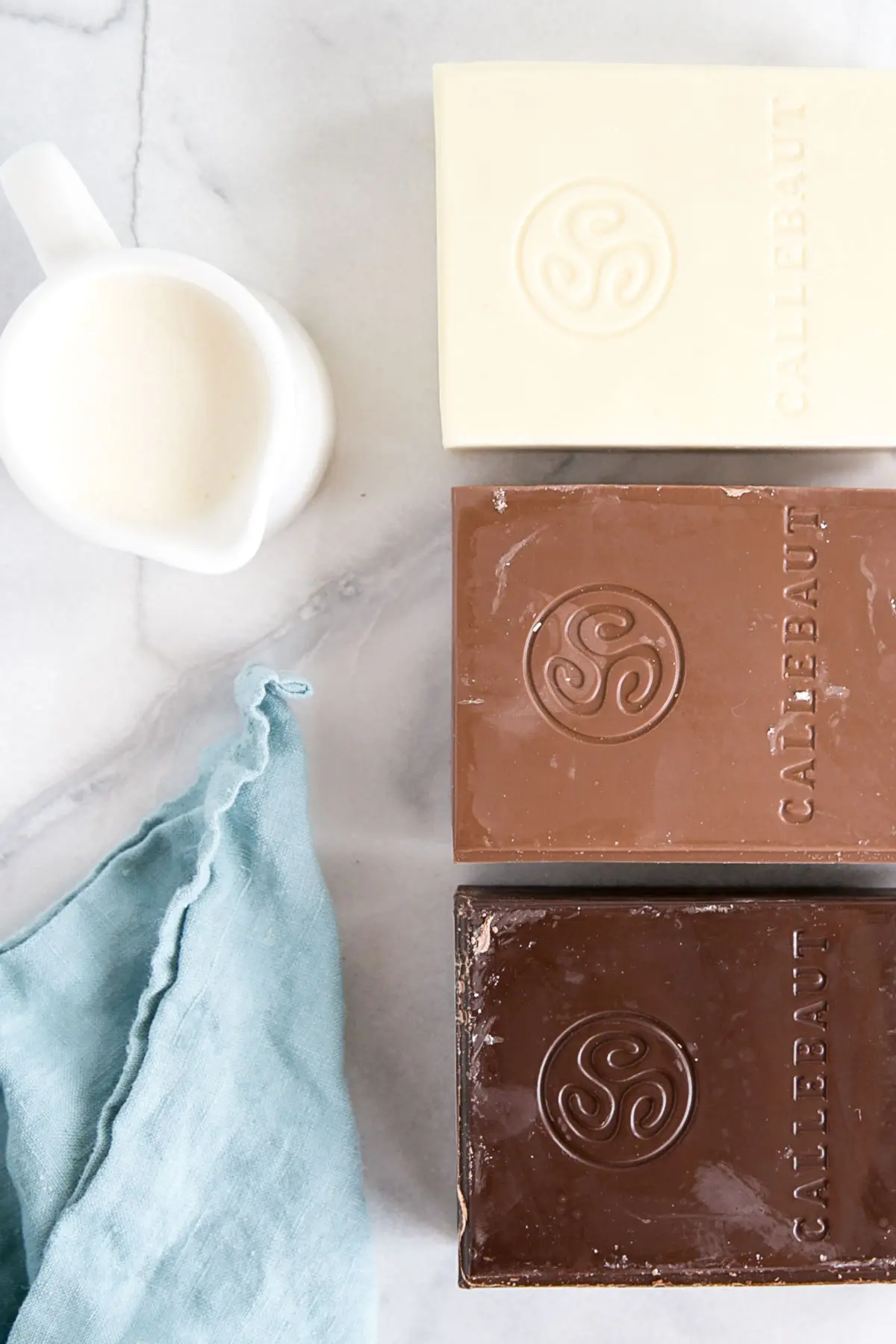 How to make Chocolate Ganache! A simple, two-ingredient recipe for a delicious frosting, filling, drip, or glaze. | livforcake.com White, milk, and dark chocolate bars beside a small pitcher of cream.