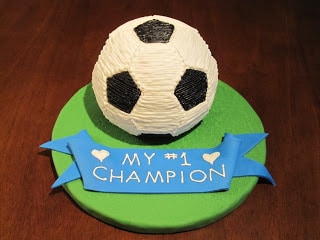 Soccer ball cake on a green cake board with a blue ribbon that says My #1 Champion.