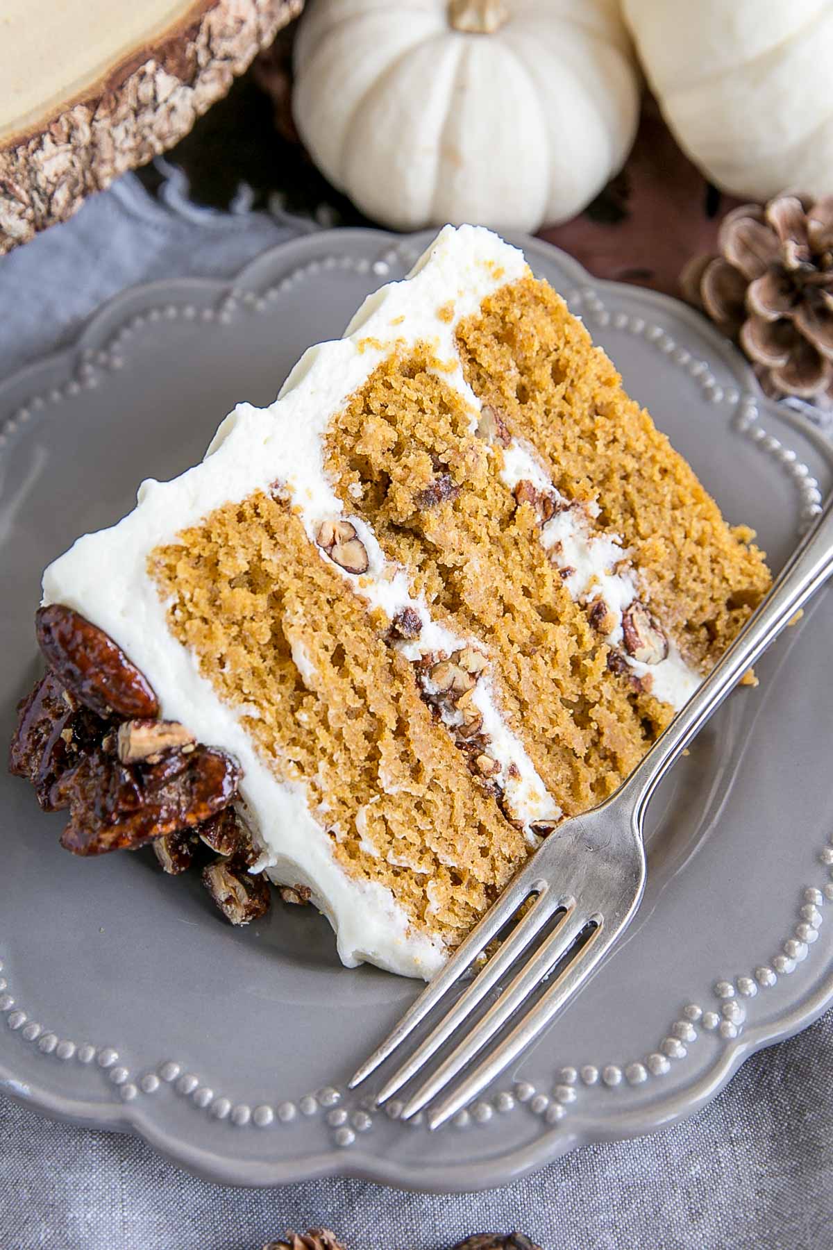 This pumpkin cake is perfect for Fall! Moist and delicious pumpkin cake layers with a tangy cream cheese frosting and crunchy candied pecans. | livforcake.com Slice of cake on a plate. Three layers of cake with candied pecans and cream cheese frosting.