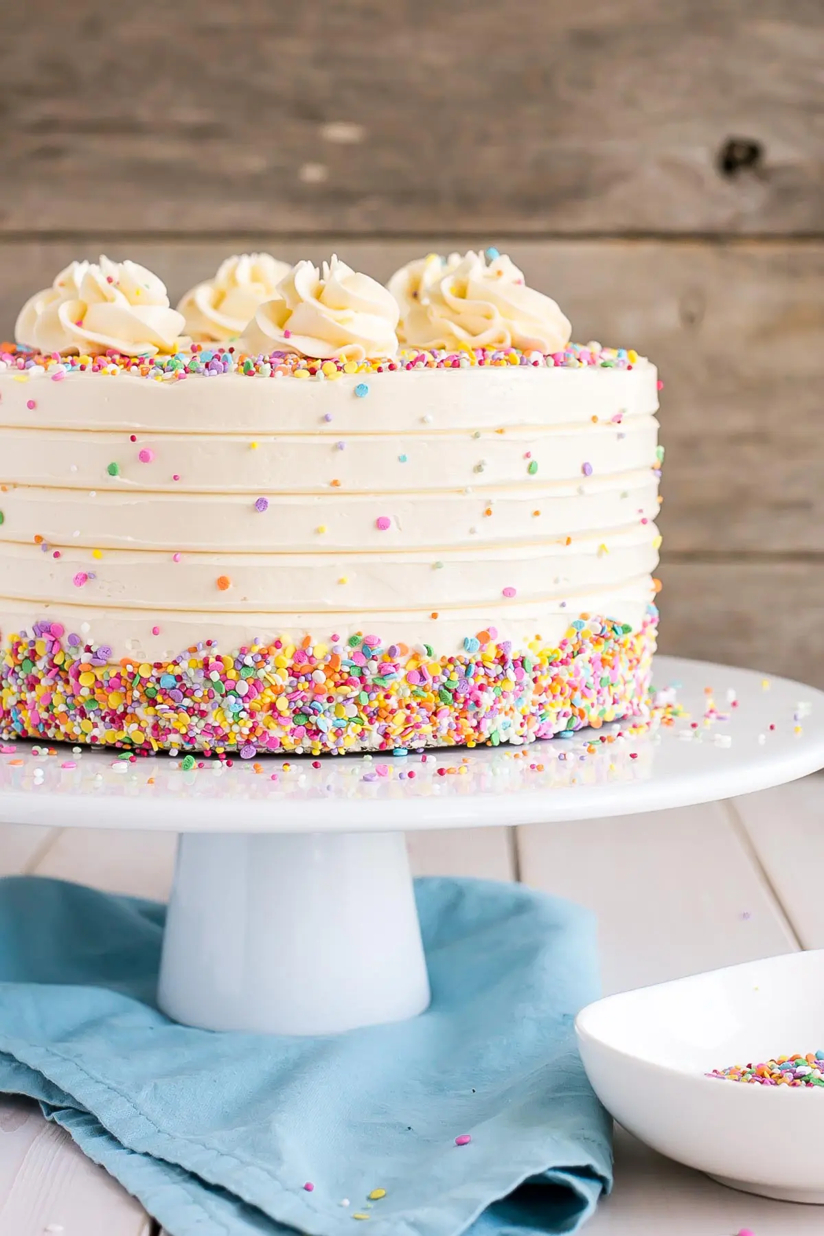 This Classic Vanilla Cake pairs fluffy vanilla cake layers with a silky vanilla buttercream. The perfect cake for birthdays, weddings, or any occasion! | livforcake.com Cake on a white cake stand, decorated with confetti sprinkles.